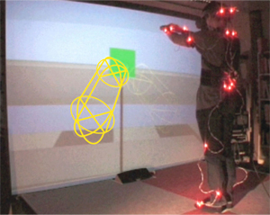 User driving simple rigid shapes at the hands in a first person viewpoint. One capsule has been enhanced for this document for visibility reasons. In this snapshot the first-person viewpoint is also displayed on the large screen for assessment purpose by an external operator.